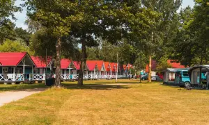 Rząd kolorowych domków letniskowych w Łebie z czerwonymi dachami, otoczonych zielenią. Obok namioty kempingowe i kamper.