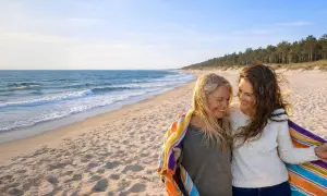 Dwie kobiety cieszą się wspólnym noclegiem w Łebie, otulone kolorowym ręcznikiem na piaszczystej plaży.