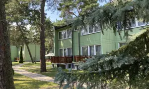 Zielony ośrodek wypoczynkowy przy wydmach, otoczony sosnami. Ścieżka prowadzi do budynków z drewnianymi tarasami.