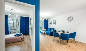 Nowoczesny pokój nad morzem z niebieskimi akcentami. Widok na salon z sofą i jadalnią oraz sypialnię z łóżkiem.
