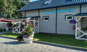 Sarbinowo domki przy plaży. Białe pergole, kwitnące kwiaty i zielony trawnik przed drewnianym budynkiem z logo.
