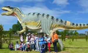 Grupa dzieci i dorosłych pozuje przed gigantycznym dinozaurem w bałtyckim parku dinozaurów.