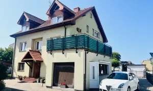 Tanie kwatery prywatne nad morzem. Dom z balkonem i białym samochodem przed posesją, otoczony zielenią.