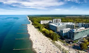 Nowoczesny hotel nad samym morzem, z widokiem na piaszczystą plażę pełną wypoczywających ludzi i turkusowe morze.