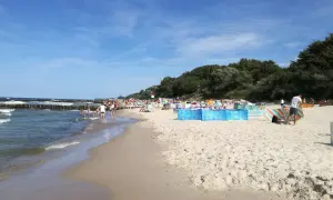 Słoneczny dzień nad morzem, plaża pełna ludzi, falochrony i zielony las w tle. Sianożęty nad morzem tętni życiem.
