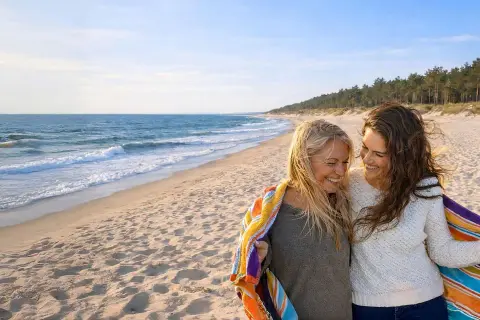 Dwie kobiety cieszą się wspólnym noclegiem w Łebie, otulone kolorowym ręcznikiem na piaszczystej plaży.