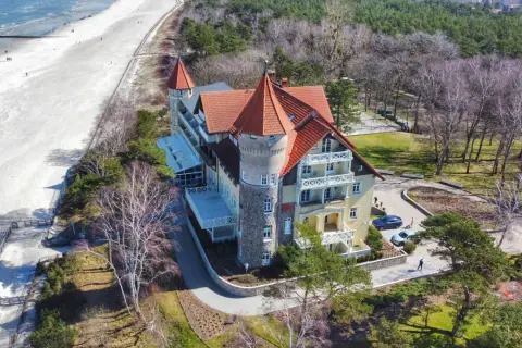 Elegancki hotel z wieżą i czerwonym dachem oferuje noclegi Łeba blisko morza. Piękna plaża i las otaczają obiekt.