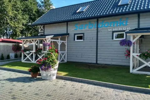 Sarbinowo domki przy plaży. Białe pergole, kwitnące kwiaty i zielony trawnik przed drewnianym budynkiem z logo.