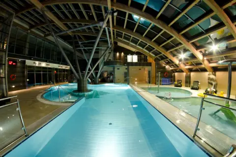 Wnętrze Laguna Kompleks Wypoczynkowo Rekreacyjny Mrzeżyno: baseny, jacuzzi, leżaki pod drewnianą, przeszkloną konstrukcją dachu.