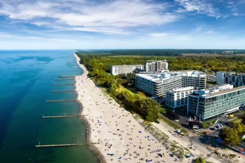 Nowoczesny hotel nad samym morzem, z widokiem na piaszczystą plażę pełną wypoczywających ludzi i turkusowe morze.