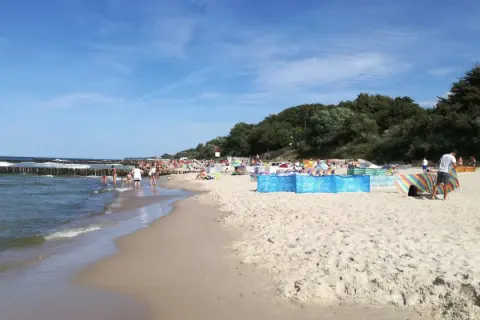 Słoneczny dzień nad morzem, plaża pełna ludzi, falochrony i zielony las w tle. Sianożęty nad morzem tętni życiem.
