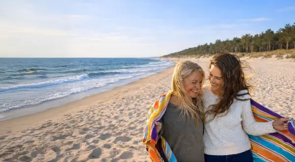 Dwie kobiety cieszą się wspólnym noclegiem w Łebie, otulone kolorowym ręcznikiem na piaszczystej plaży.