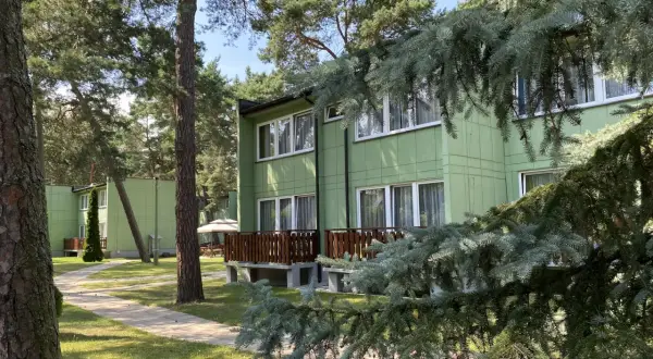 Zielony ośrodek wypoczynkowy przy wydmach, otoczony sosnami. Ścieżka prowadzi do budynków z drewnianymi tarasami.