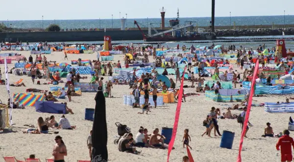 Tłum ludzi na plaży w Łebie. W tle widać morze i port. Tyle mieszkańców ma Łeba, że plaża jest tak pełna.