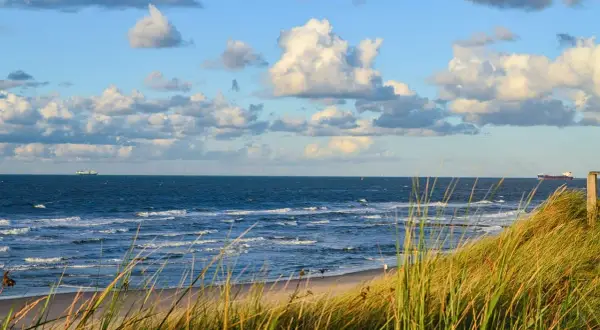 Piękny widok na morze z piaszczystą plażą i wydmami porośniętymi trawą. Na horyzoncie widać statki, a niebo zdobią chmury. To idealne miejsce na spacer, gdzie można poczuć morską bryzę i podziwiać krajobraz.