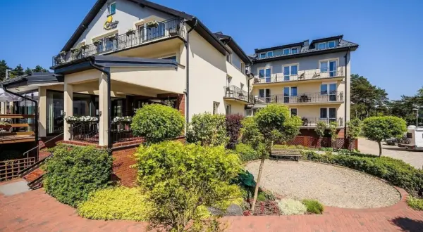 Hotel La Siesta w słoneczny dzień. Budynek z balkonami otoczony zielenią i brukowanym dziedzińcem.