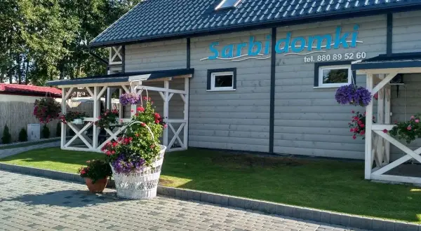 Sarbinowo domki przy plaży. Białe pergole, kwitnące kwiaty i zielony trawnik przed drewnianym budynkiem z logo.