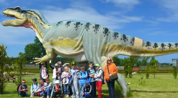 Grupa dzieci i dorosłych pozuje przed gigantycznym dinozaurem w bałtyckim parku dinozaurów.