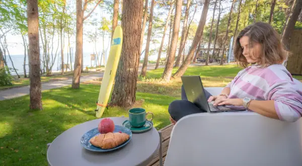 Kobieta pracuje na laptopie przy stoliku z kawą i croissantem. W tle widać morze i domki z wyjściem na plażę.