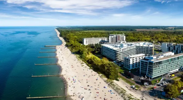 Nowoczesny hotel nad samym morzem, z widokiem na piaszczystą plażę pełną wypoczywających ludzi i turkusowe morze.