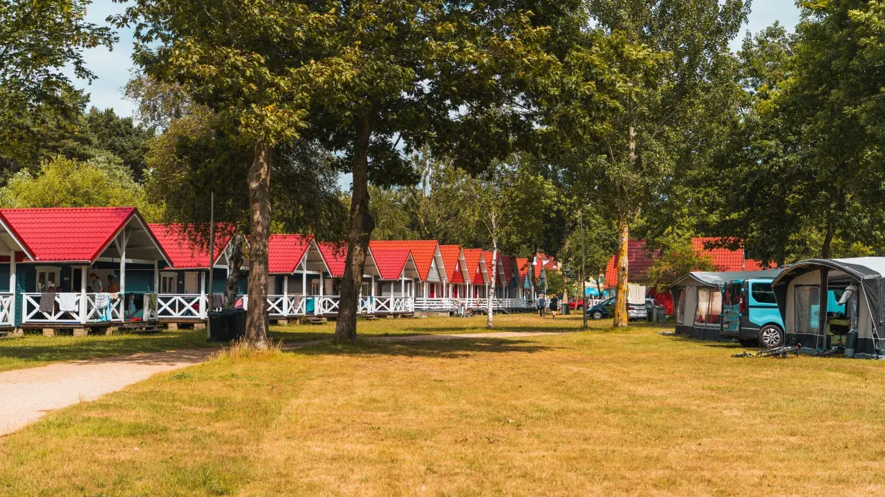 Rząd kolorowych domków letniskowych w Łebie z czerwonymi dachami, otoczonych zielenią. Obok namioty kempingowe i kamper.