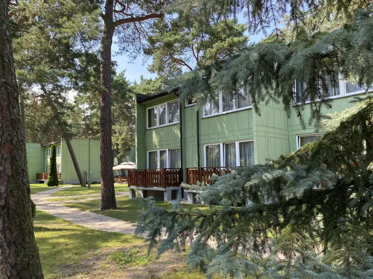 Zielony ośrodek wypoczynkowy przy wydmach, otoczony sosnami. Ścieżka prowadzi do budynków z drewnianymi tarasami.