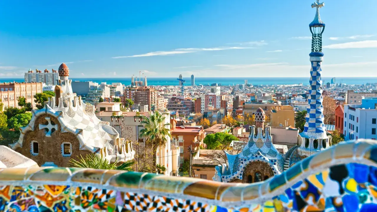 Widok na kolorowe budowle Parku Güell i panoramę miasta, gdzie leży Barcelona, z morzem w tle.