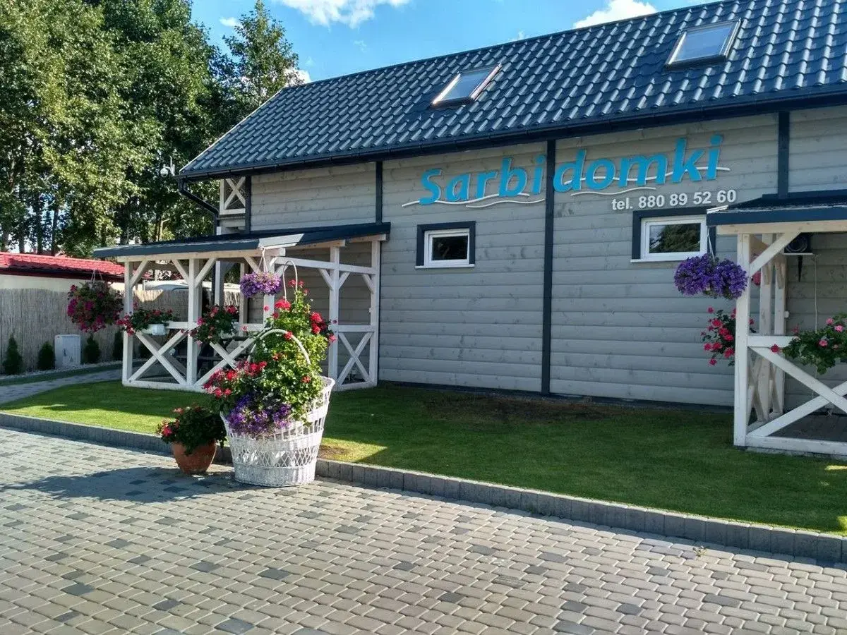 Sarbinowo domki przy plaży. Białe pergole, kwitnące kwiaty i zielony trawnik przed drewnianym budynkiem z logo.