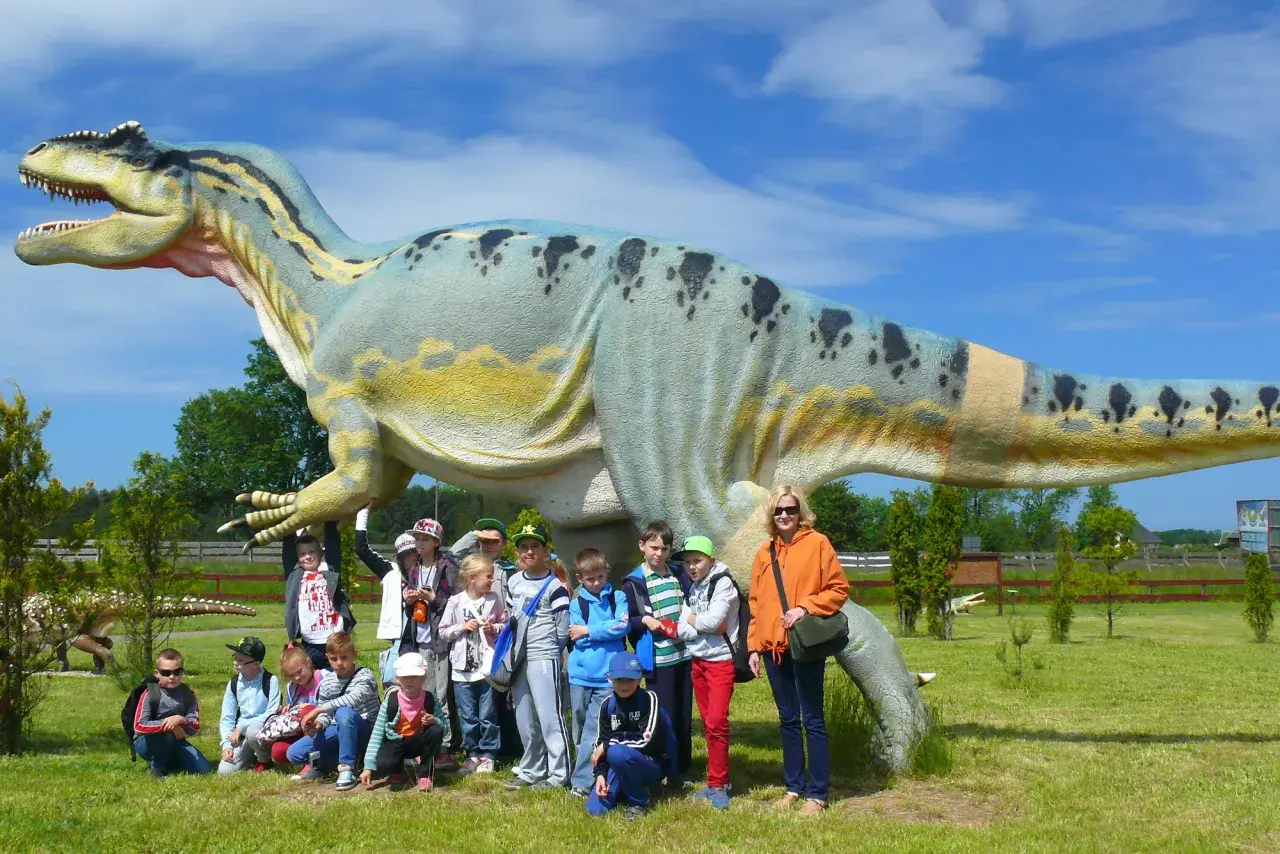 Grupa dzieci i dorosłych pozuje przed gigantycznym dinozaurem w bałtyckim parku dinozaurów.