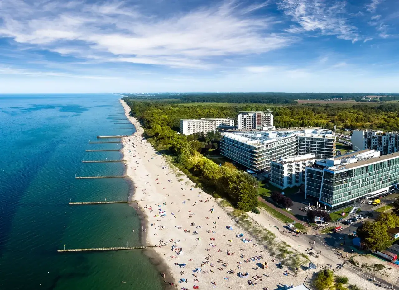 Nowoczesny hotel nad samym morzem, z widokiem na piaszczystą plażę pełną wypoczywających ludzi i turkusowe morze.