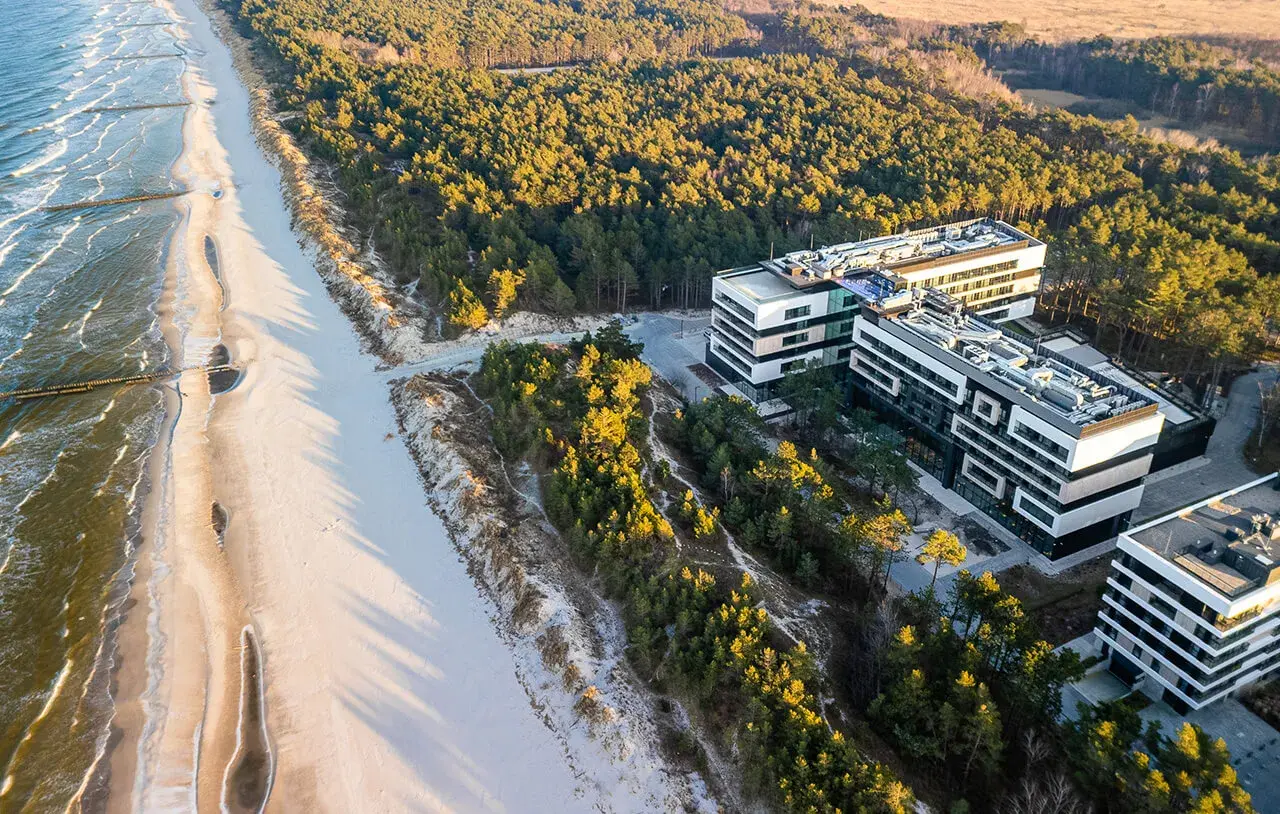 Nowoczesne budynki hotelowe w Rogowie nad morzem, otoczone sosnowym lasem, z widokiem na piaszczystą plażę i fale Bałtyku.