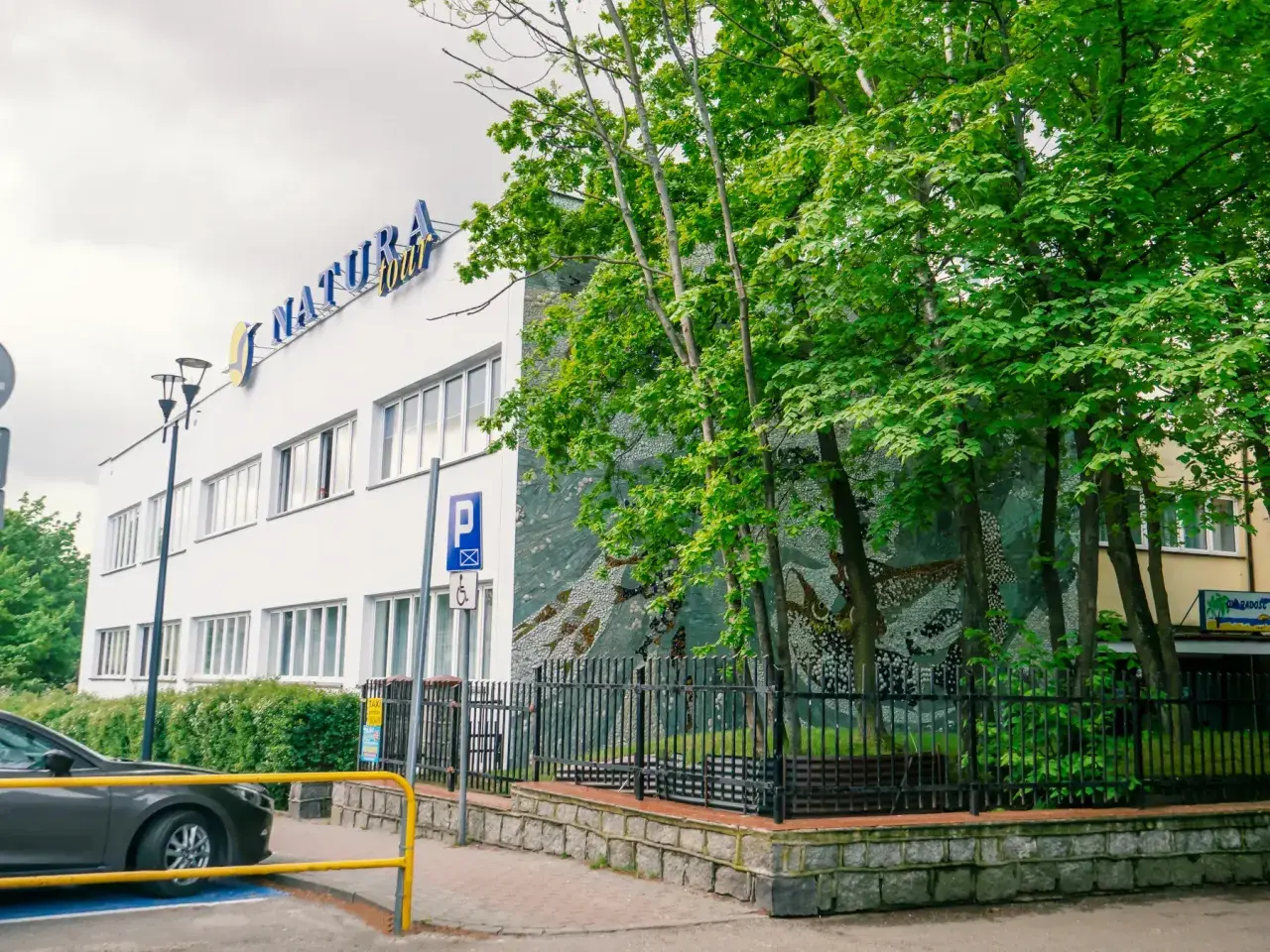 Dom wczasowy Radość, budynek z niebieskim napisem "NATURA tour", otoczony zielenią i ozdobiony mozaiką.