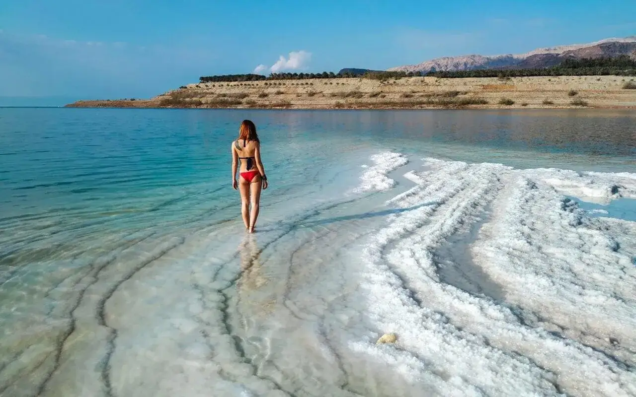 Kobieta w czerwonym bikini spaceruje po krystalicznej plaży Morza Martwego. Nie ma tu rekinów, tylko niezwykłe krajobrazy.