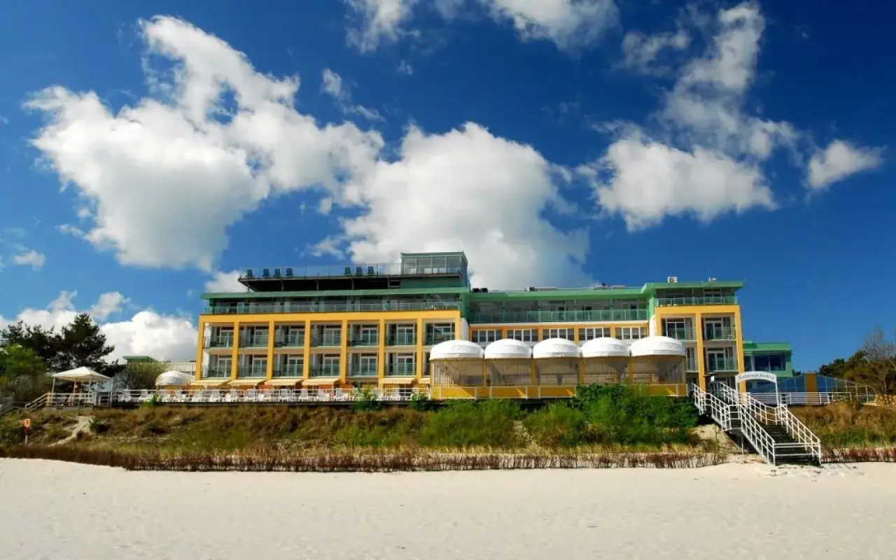 Jurata noclegi z wyżywieniem: nowoczesny hotel z żółtą fasadą, tarasami i widokiem na plażę i morze.