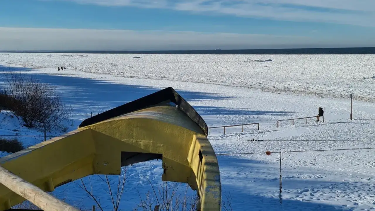 Zimowy krajobraz nad morzem, gdzie widać, czy morze zamarza. Na pierwszym planie żółta łódź, w oddali ludzie spacerujący po lodzie.