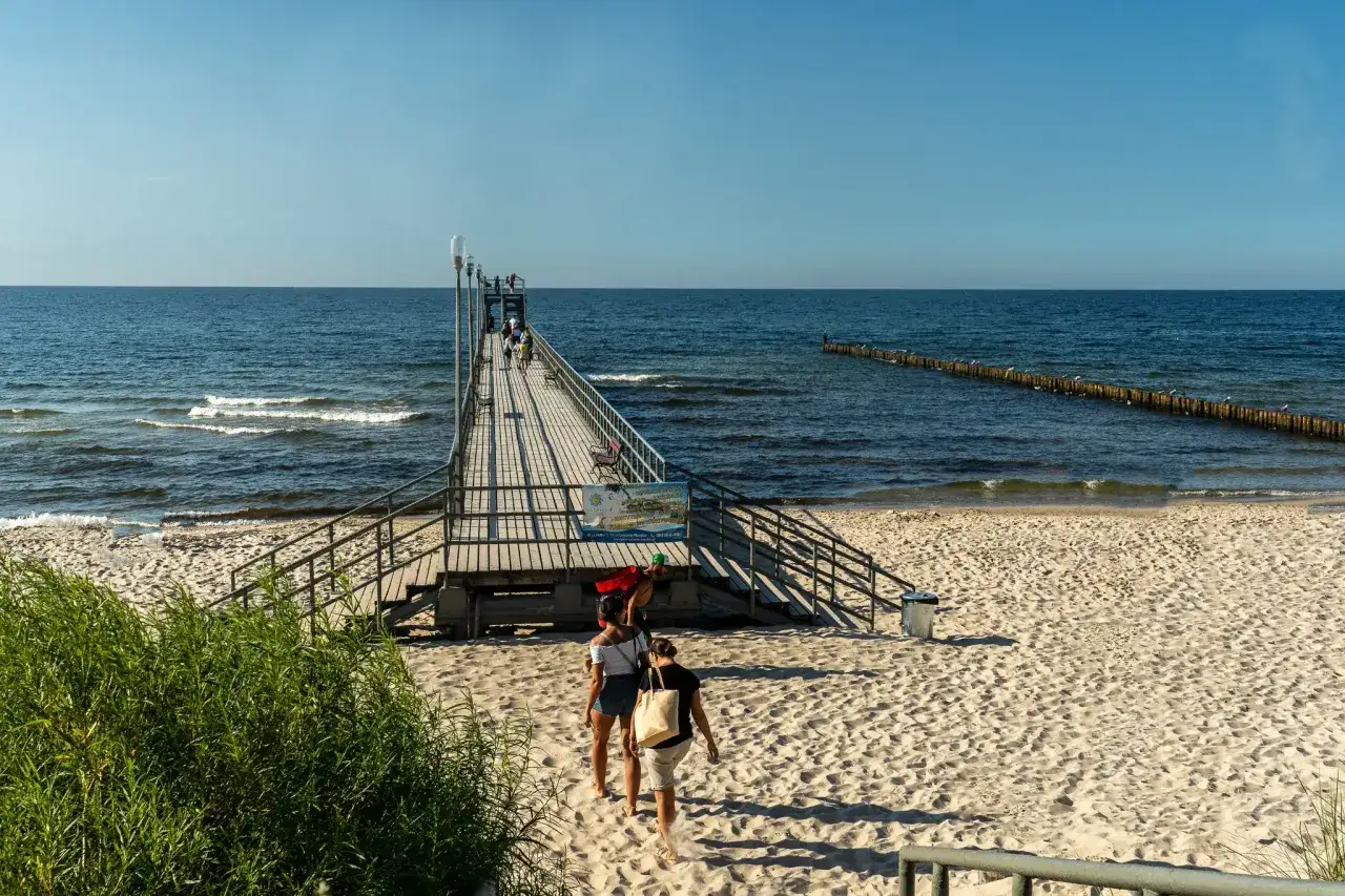Sianożęty nad morzem: drewniane molo wchodzi w głąb spokojnego, błękitnego morza, a na plaży widać spacerujących ludzi.