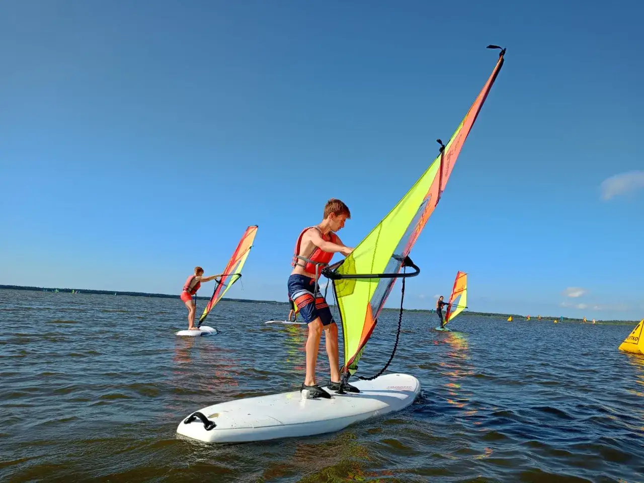 Chłopcy uczą się windsurfingu na jeziorze, niedaleko plaży w Dąbkach. Słoneczny dzień sprzyja wodnym szaleństwom.