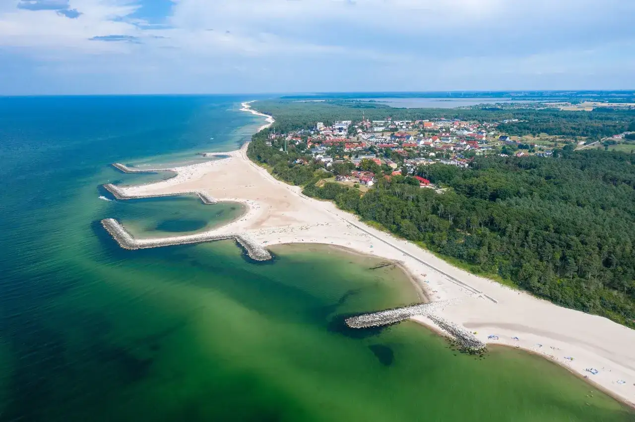 Malownicze wybrzeże bałtyckie miejscowości z kamiennymi falochronami, piaszczystą plażą i zielonym lasem w tle.