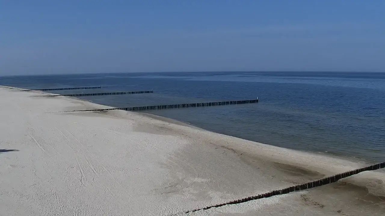 Pusta plaża w Rogowie nad morzem, z drewnianymi falochronami wchodzącymi w spokojne, błękitne morze.