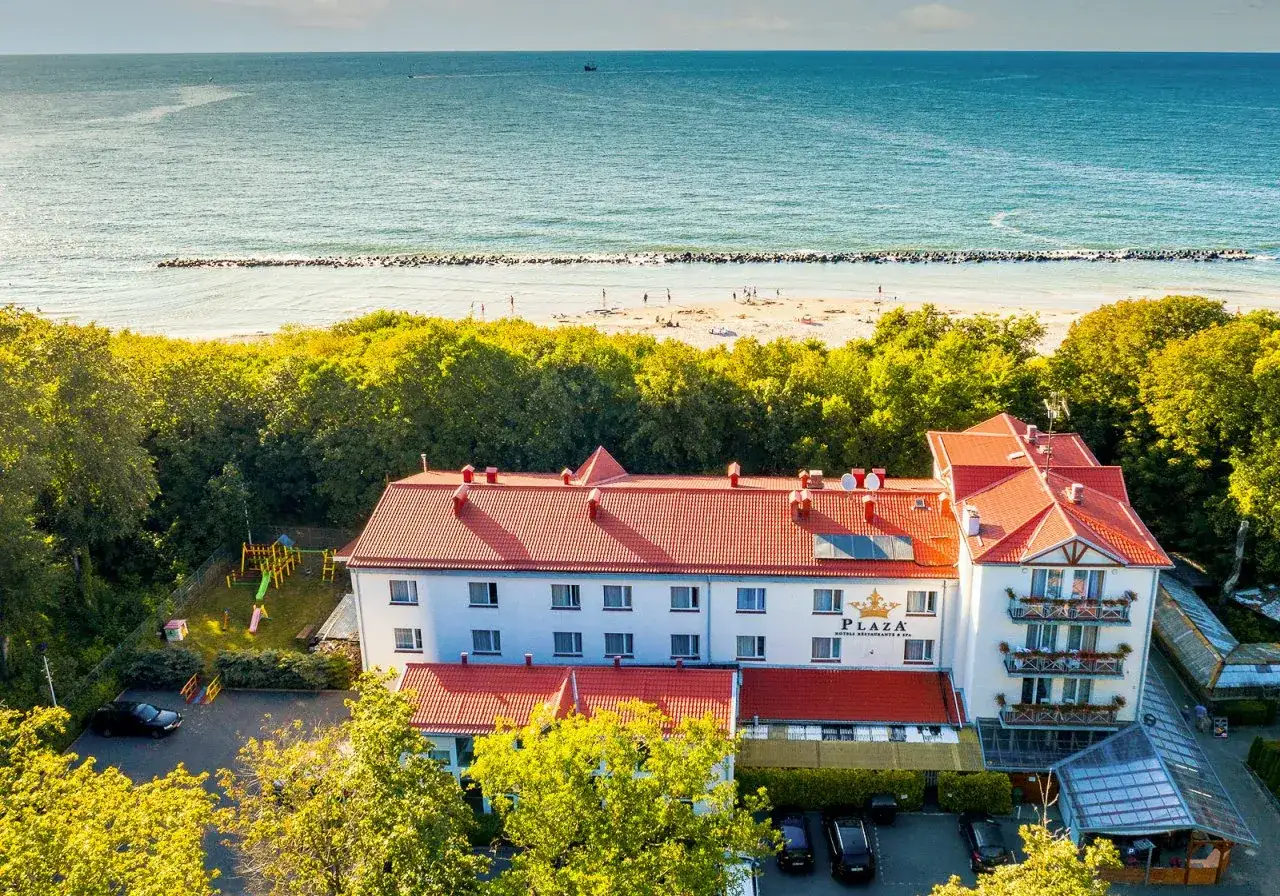 Ośrodek wczasowy Słowianka tuż przy plaży. Biały budynek z czerwonym dachem, otoczony zielenią, z widokiem na morze.