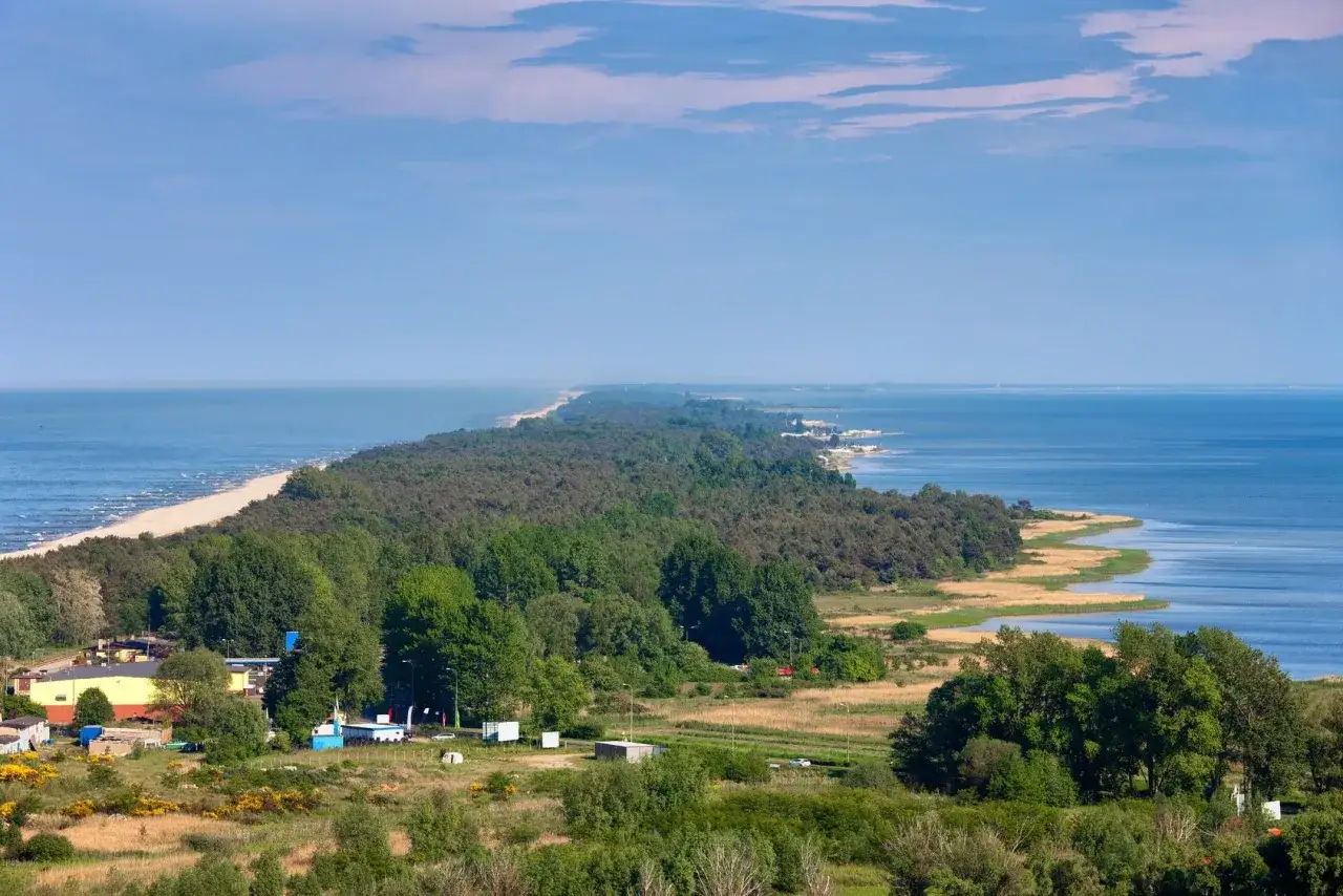 Ośrodek wypoczynkowy przy wydmach, z widokiem na morze i las. Długi, piaszczysty brzeg plaży ciągnie się w oddali.