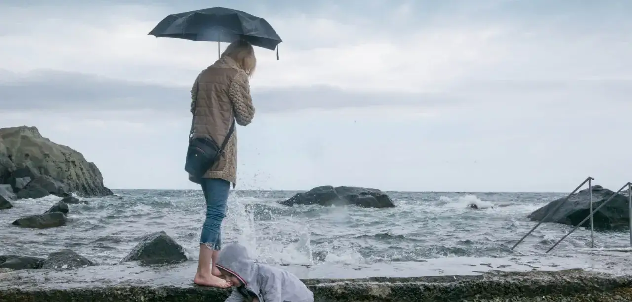 Dzieci bawią się na kamienistym brzegu podczas wycieczki nad morze. Fale rozbijają się o brzeg, a jedna z fal obryzguje dziecko.