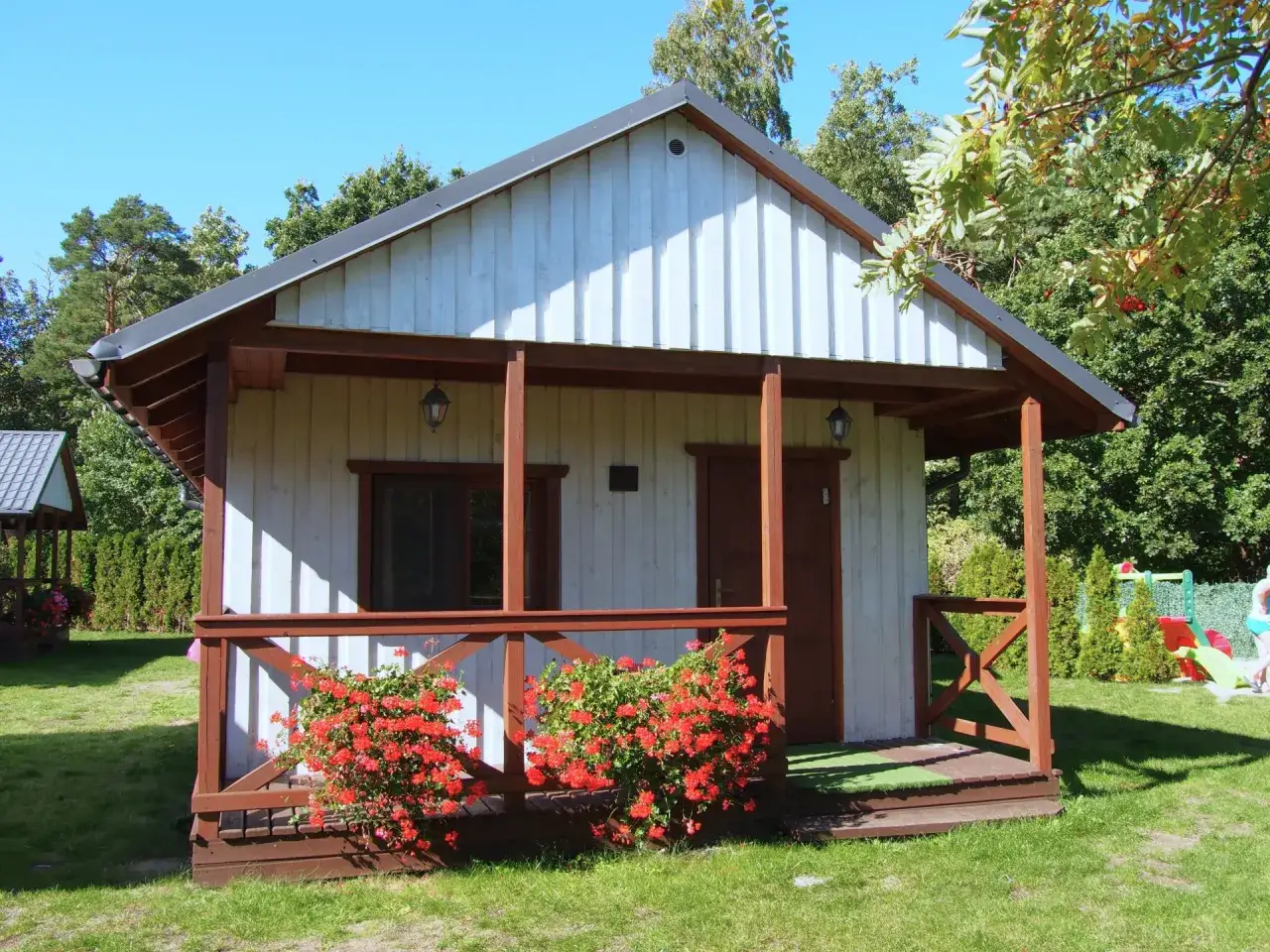Urocze domki letniskowe Amber House w otoczeniu zieleni. Przed domkiem kwitną czerwone pelargonie.
