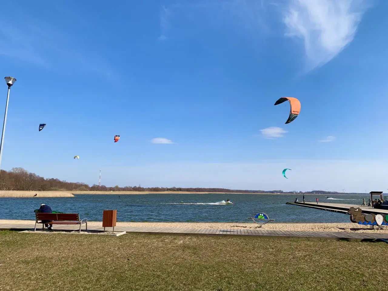 Wietrzny dzień nad jeziorem w Dąbkach, gdzie kitesurferzy ścigają się na falach. W oddali widać dom przy plaży.