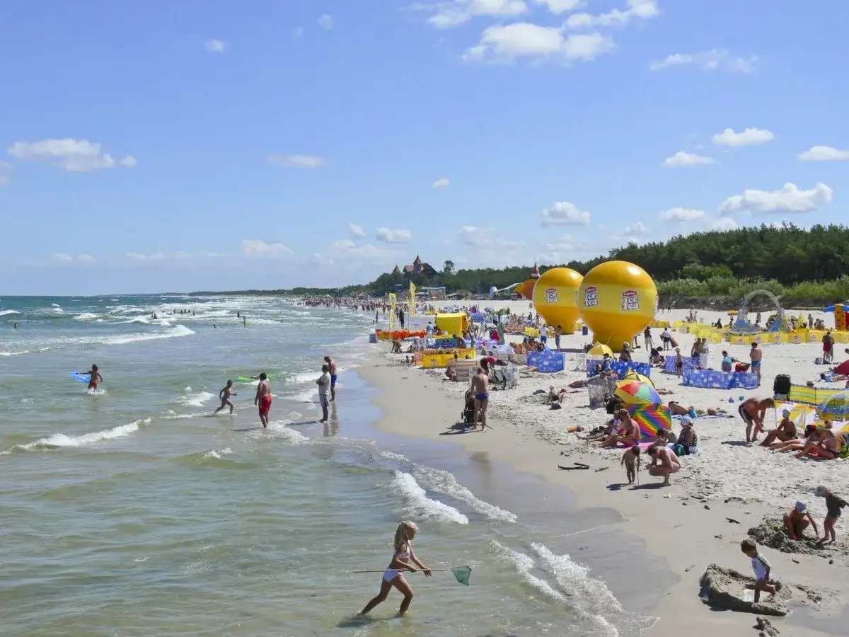 Słoneczna plaża w Łebie, pełna ludzi, z falami i dmuchanymi balonami. Idealne miejsce na łeba noclegi z wyżywieniem.