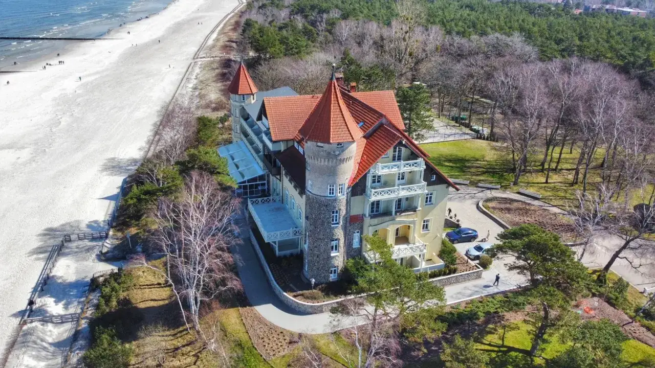 Elegancki hotel z wieżą i czerwonym dachem, idealny na noclegi Łeba blisko morza. Piękna plaża i las otaczają budynek.