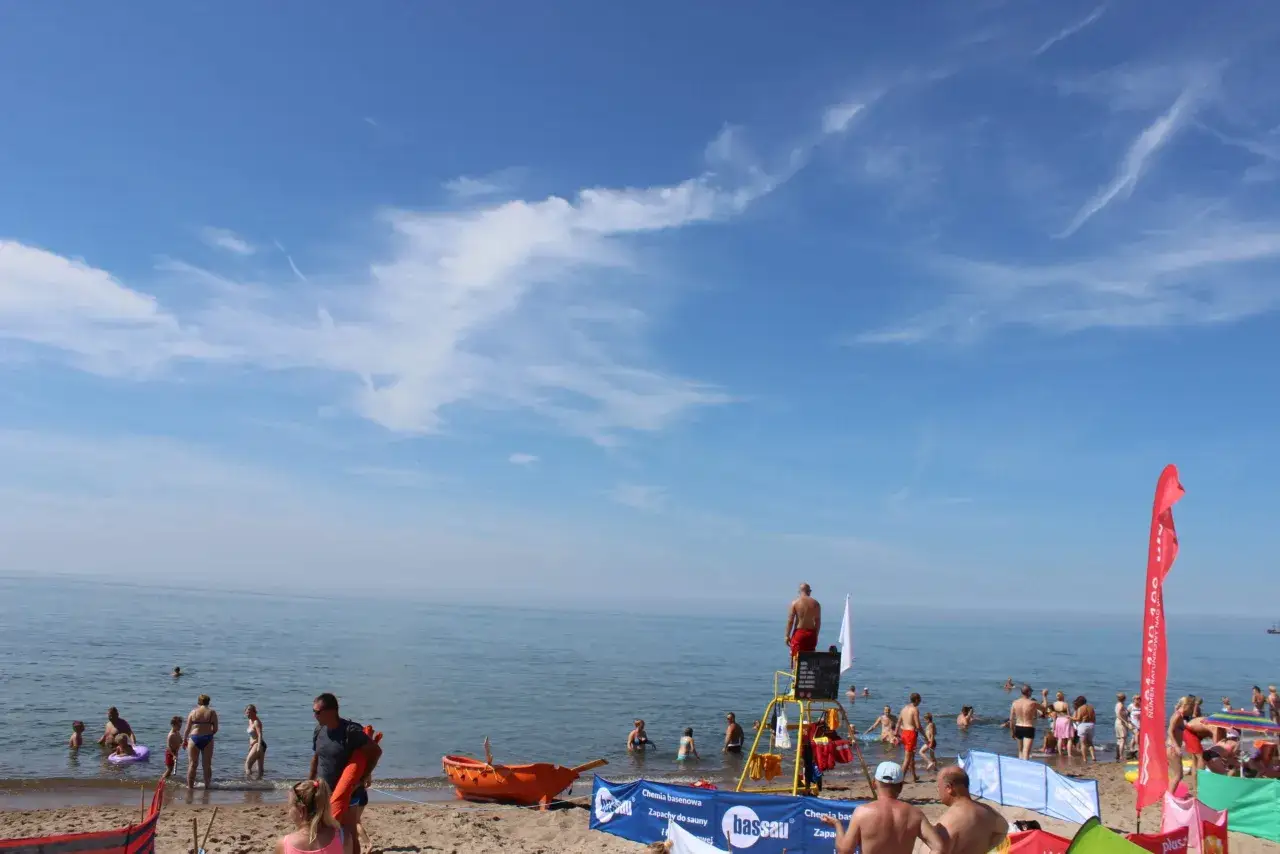 Słoneczny wyjazd nad morze. Ludzie kąpią się w morzu, ratownik czuwa na wieży, a na plaży widać pomarańczową łódź ratunkową.