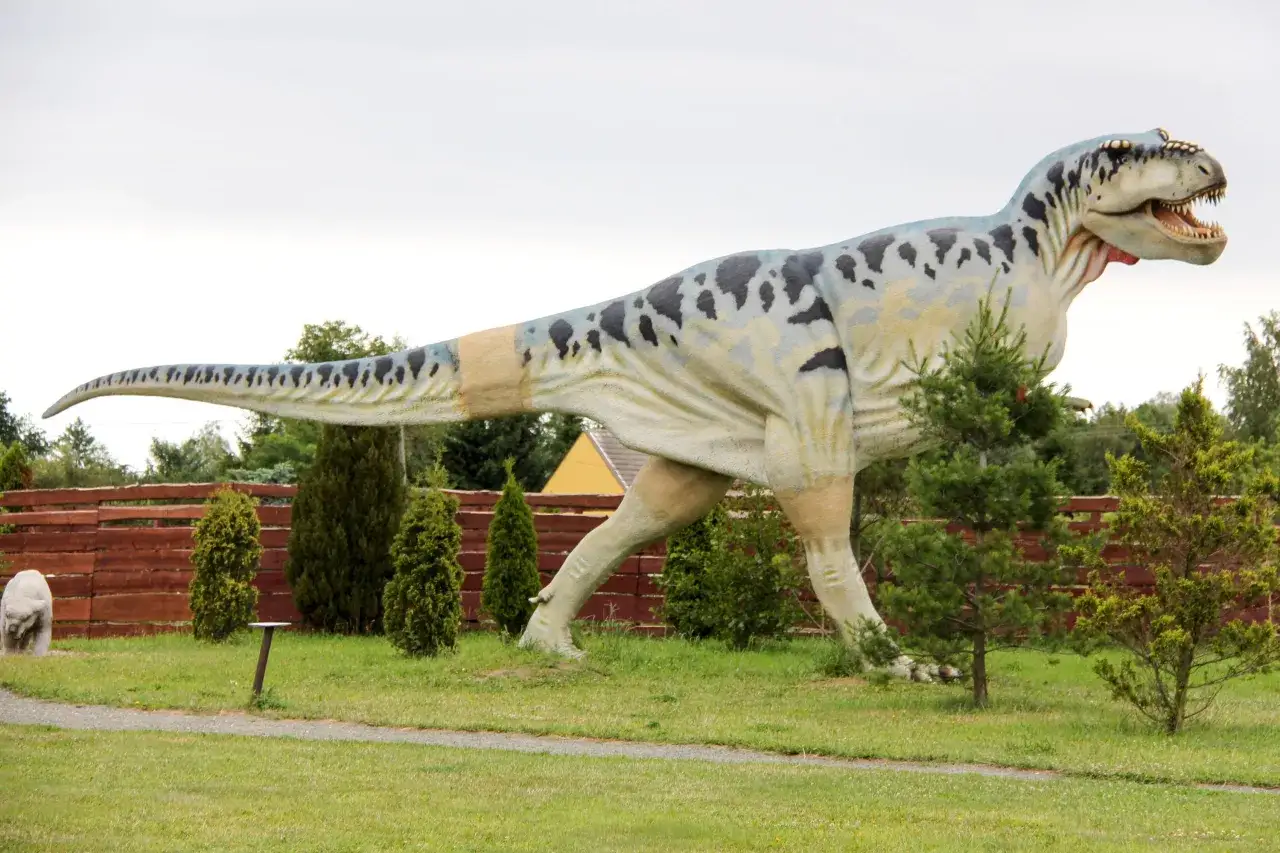 Gigantyczny model tyranozaura w bałtyckim parku dinozaurów, z plamistym wzorem na skórze, stoi na trawiastym terenie wśród drzew.