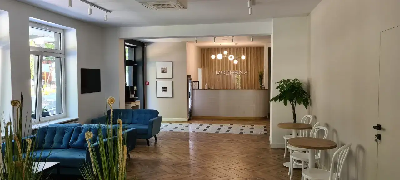 Wnętrze hotelu Moderna Jastrzębia Góra: wygodne kanapy, recepcja z drewnianą ścianą i logo, stoliki z krzesłami.