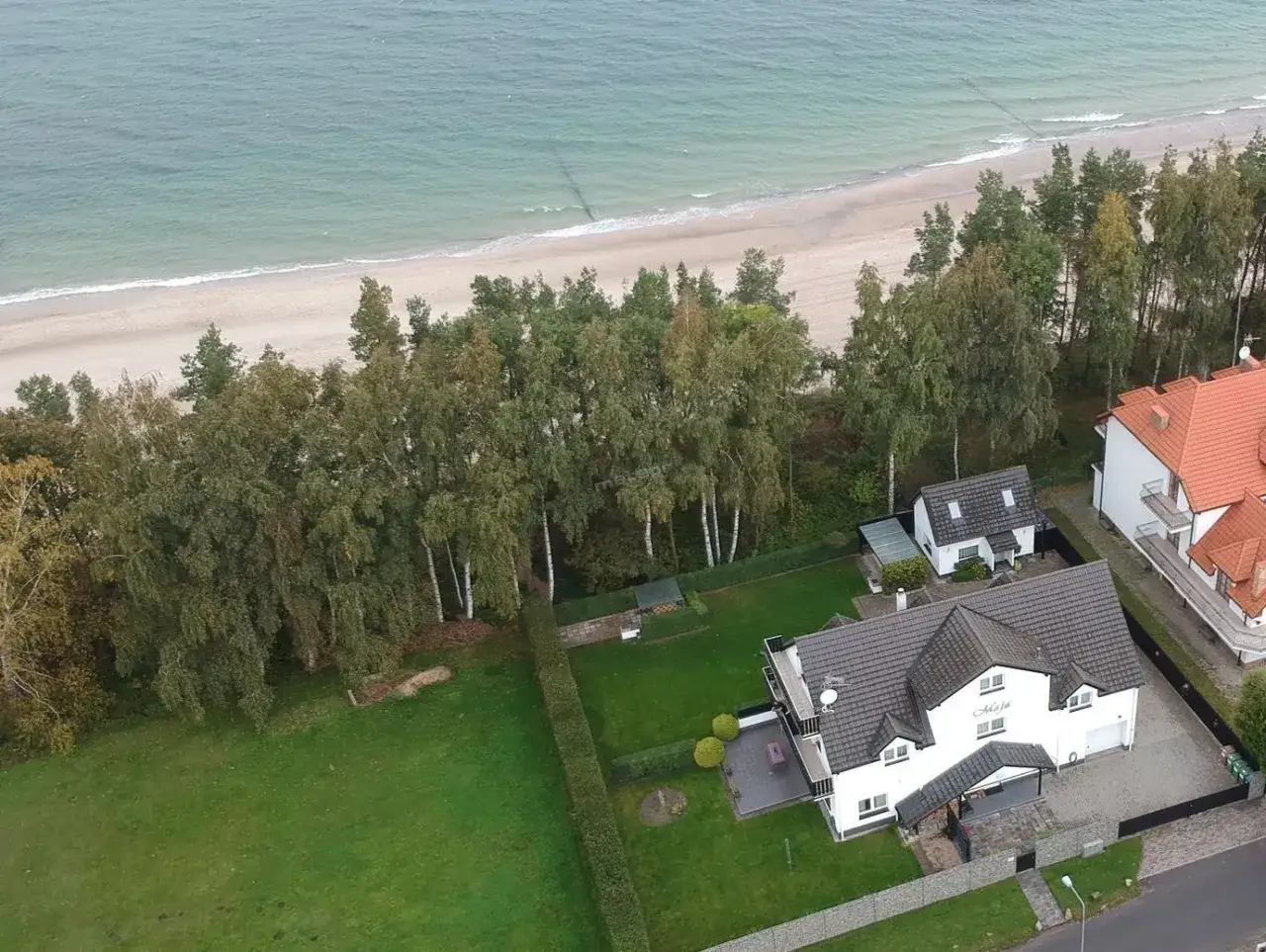 Sarbinowo domki przy plaży. Dwa białe domy z szarymi dachami, otoczone zielenią, blisko piaszczystej plaży i morza.